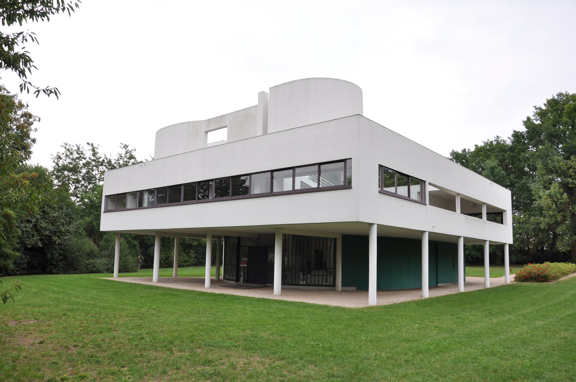 Photograph of a modernist architectural building elevated on white stilts with a flat roof and horizontal windows wrapping around the upper level. Surrounded by green grass and trees, the structure features clean white walls and an open ground floor area.
