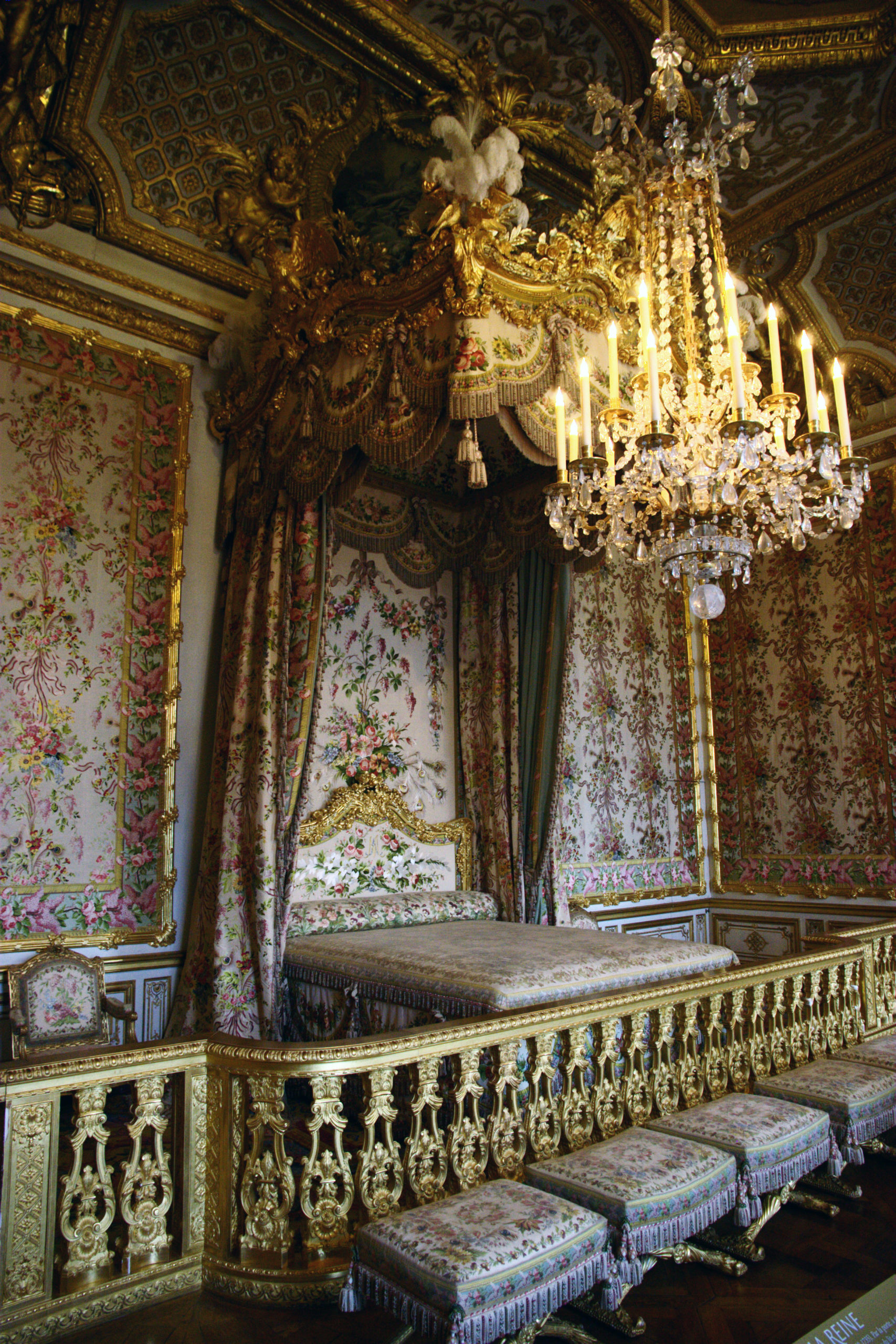 Photograph of an Queen's bed chamber, Versailles featuring a gilded canopy bed with floral upholstery and intricate gold detailing. A large crystal chandelier hangs above, illuminating richly decorated walls with floral tapestries and a matching set of upholstered stools.