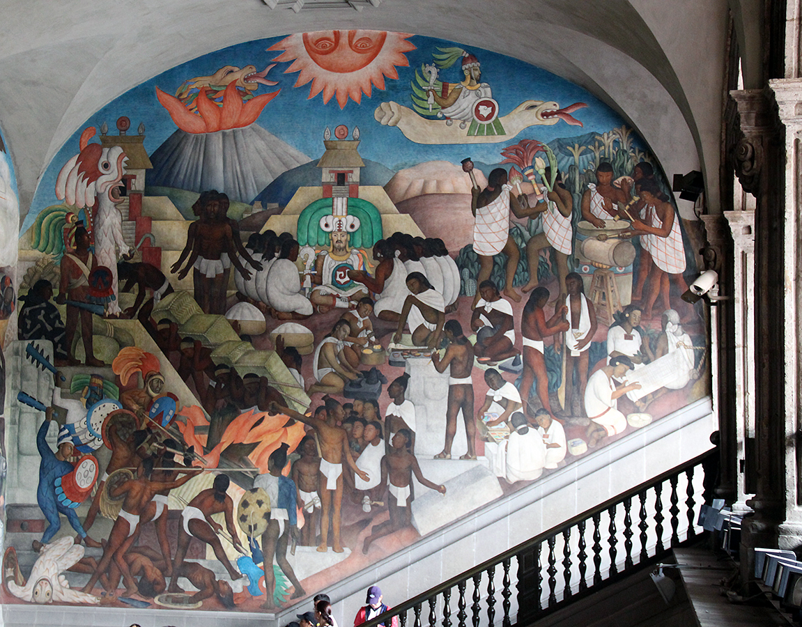 A large mural painting depicting an ancient Mesoamerican scene with numerous figures engaged in various activities such as rituals, music, and daily tasks, set against a backdrop of mountains, temples, and symbolic elements like a sun and serpent. The mural features vibrant colors, detailed human forms, and cultural motifs, illustrating historical and mythological aspects of indigenous life.