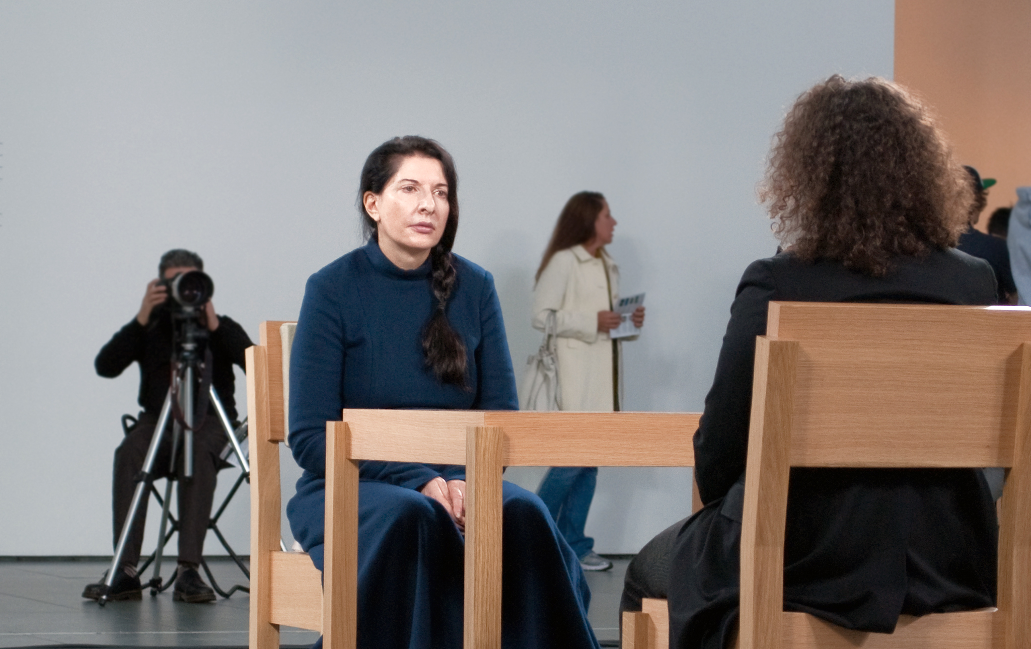 Photograph showing two women seated on wooden chairs facing each other, engaged in conversation or interview. A photographer with a camera on a tripod and a woman walking in the background are also visible, indicating a staged setting.