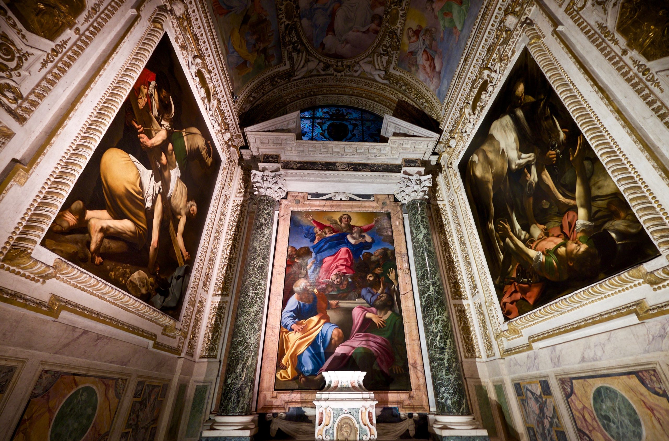 Photograph of an ornate chapel interior featuring three large religious paintings framed by intricate marble and gold architectural details. Central painting depicts a seated figure surrounded by disciples, while side paintings show dramatic biblical scenes with vivid colors and dynamic compositions.