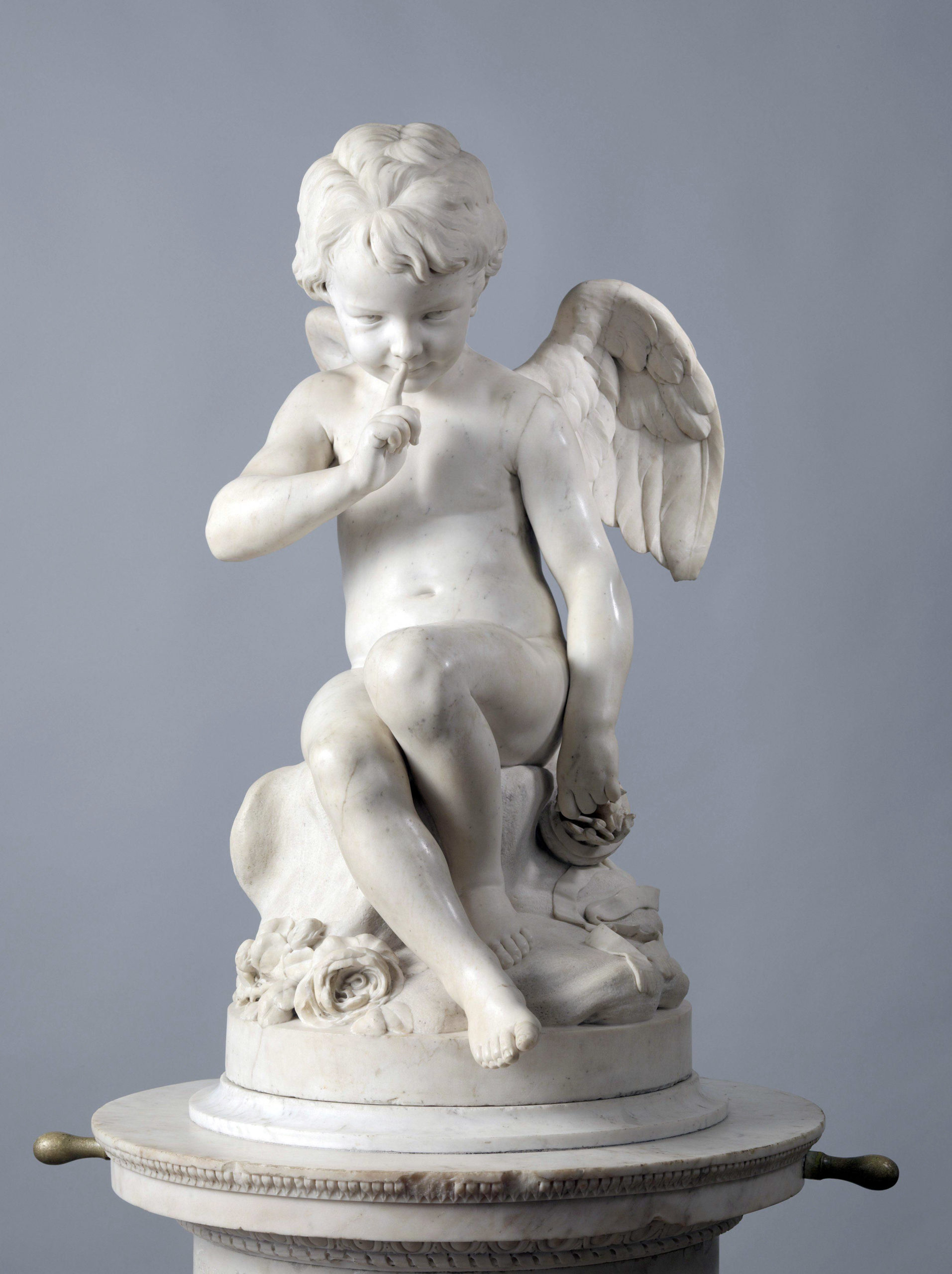 A photograph of a white marble sculpture depicting a seated cherub with detailed wings, curly hair, and a relaxed pose. The cherub rests on a round pedestal adorned with floral carvings, set against a plain gray background.