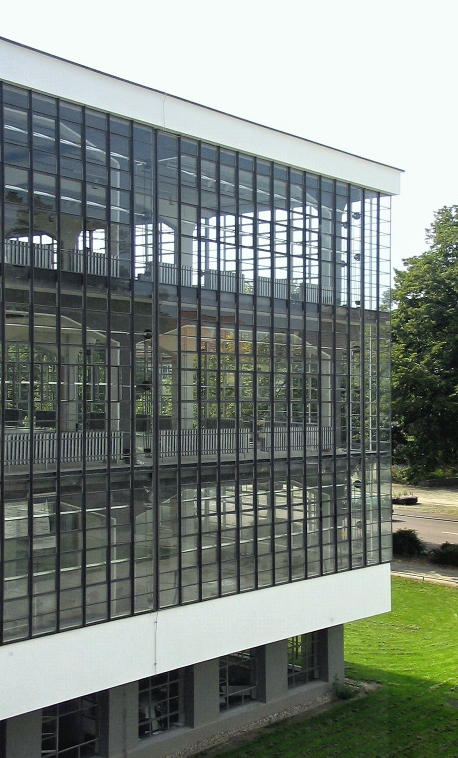 Photograph of a modern building corner featuring large glass windows with black grid frames reflecting interior structural elements and outdoor greenery. The building's white exterior contrasts with the green lawn and trees surrounding it, highlighting architectural transparency and integration with nature.