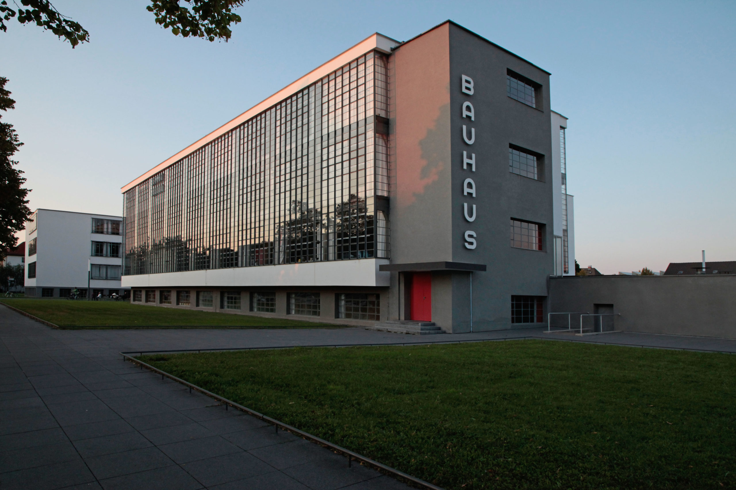 Photograph of Bauhaus school building showcasing modernist architecture with large grid-patterned windows and minimalist design. Gray facade features vertical "BAUHAUS" lettering, with red door providing color contrast, set against a clear evening sky.