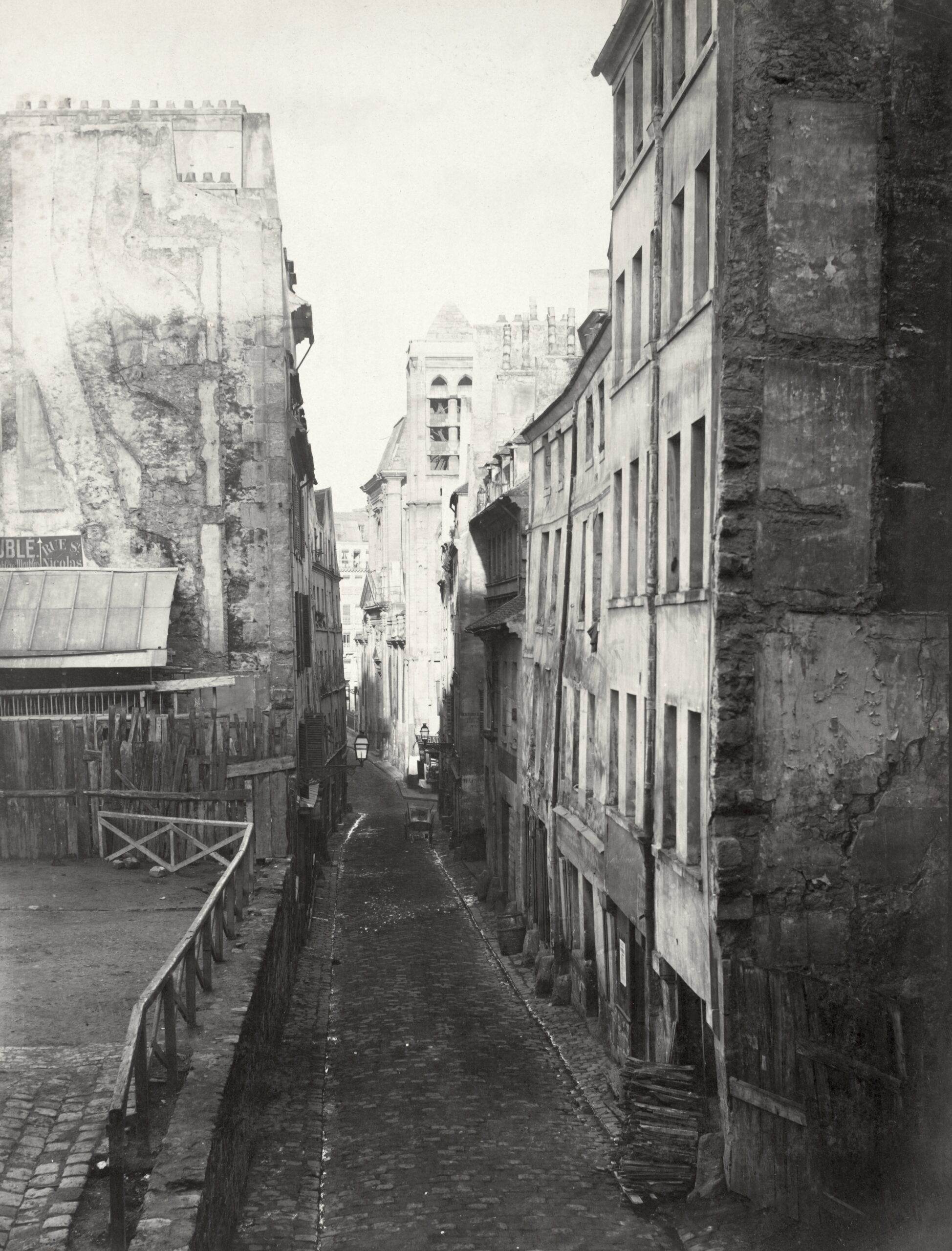 Black and white photograph of a narrow, empty street lined with tall, aged buildings showing signs of wear and decay. The scene captures an urban environment with uneven pavement, visible architectural details, and a distant view of more structures under soft daylight.
