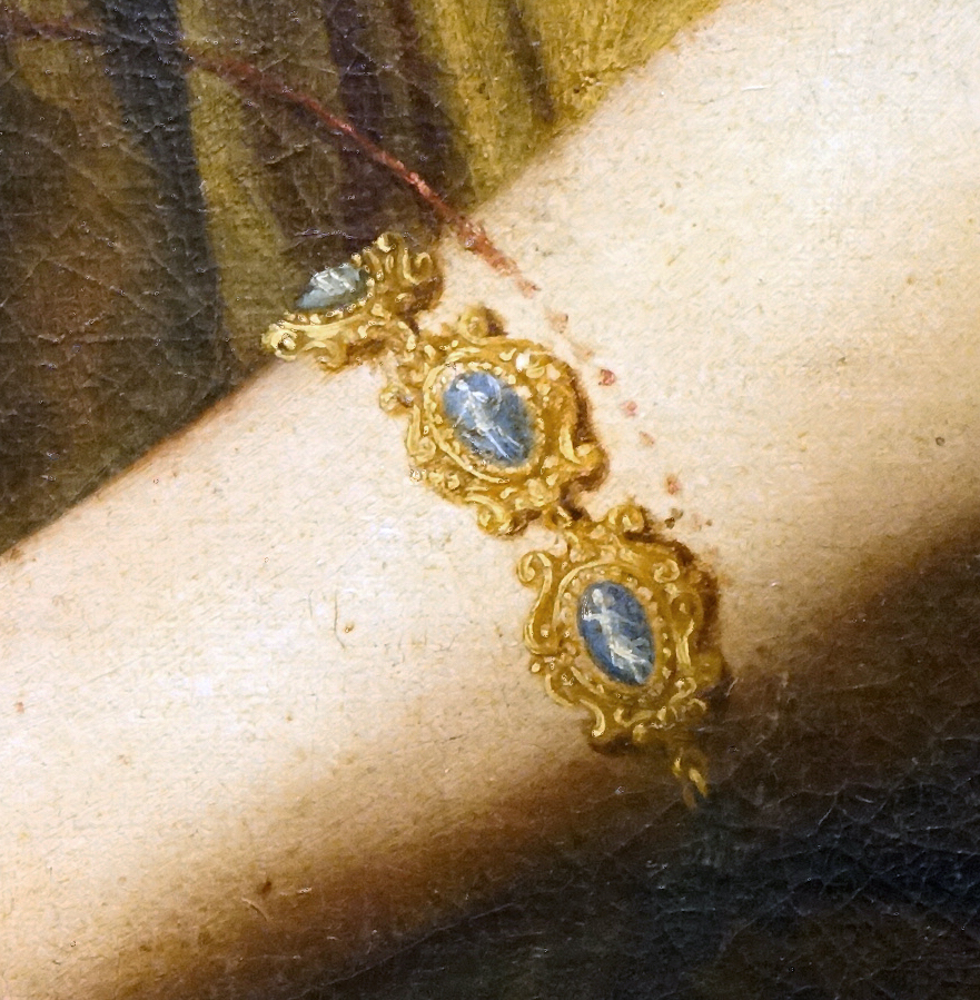 Close-up photograph of a painted arm wearing an ornate gold bracelet with three blue oval gemstones set in intricate gold frames.
