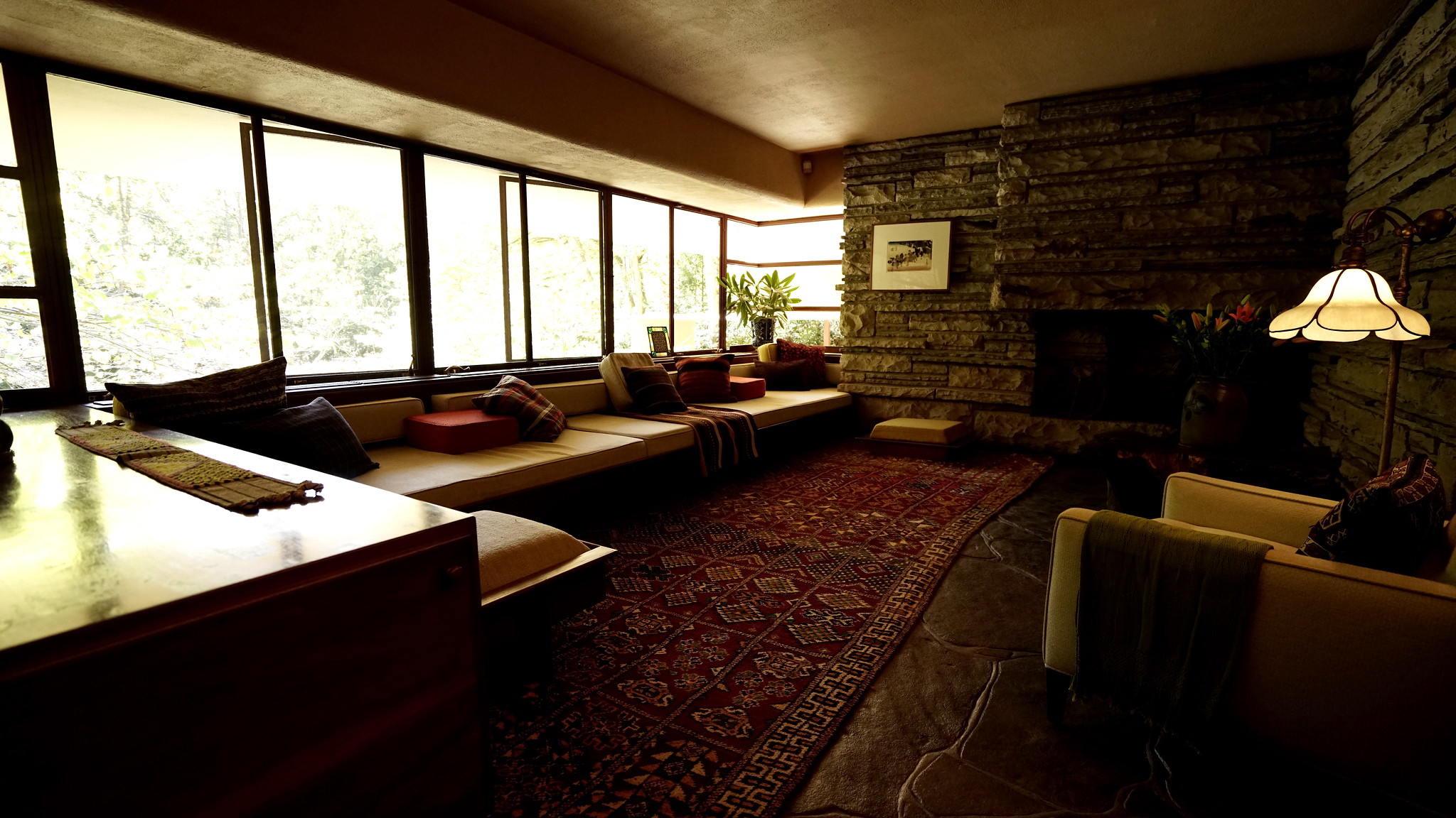 Photograph of a cozy living room featuring large windows along one wall, a long built-in bench with cushions, and a stone fireplace on the opposite side. The room includes a patterned red rug, a wooden table, an armchair with a blanket, and warm lighting from a table lamp.