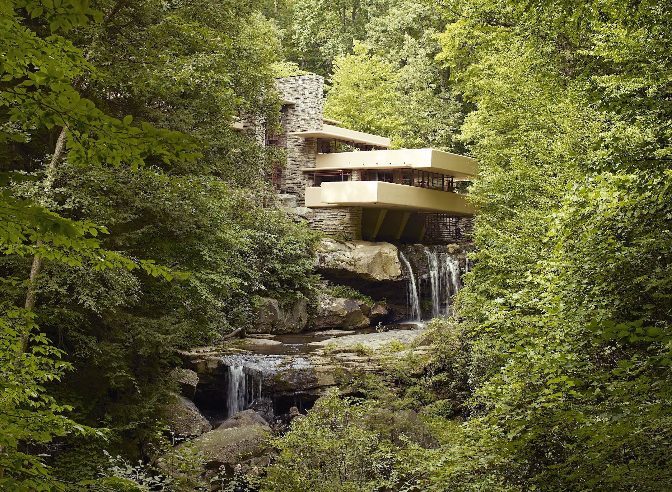 Photograph of Fallingwater, a famous house designed by Frank Lloyd Wright, integrated into a forested landscape with a waterfall flowing beneath it. The structure features cantilevered terraces in beige and stone materials, blending modern architecture with natural surroundings.