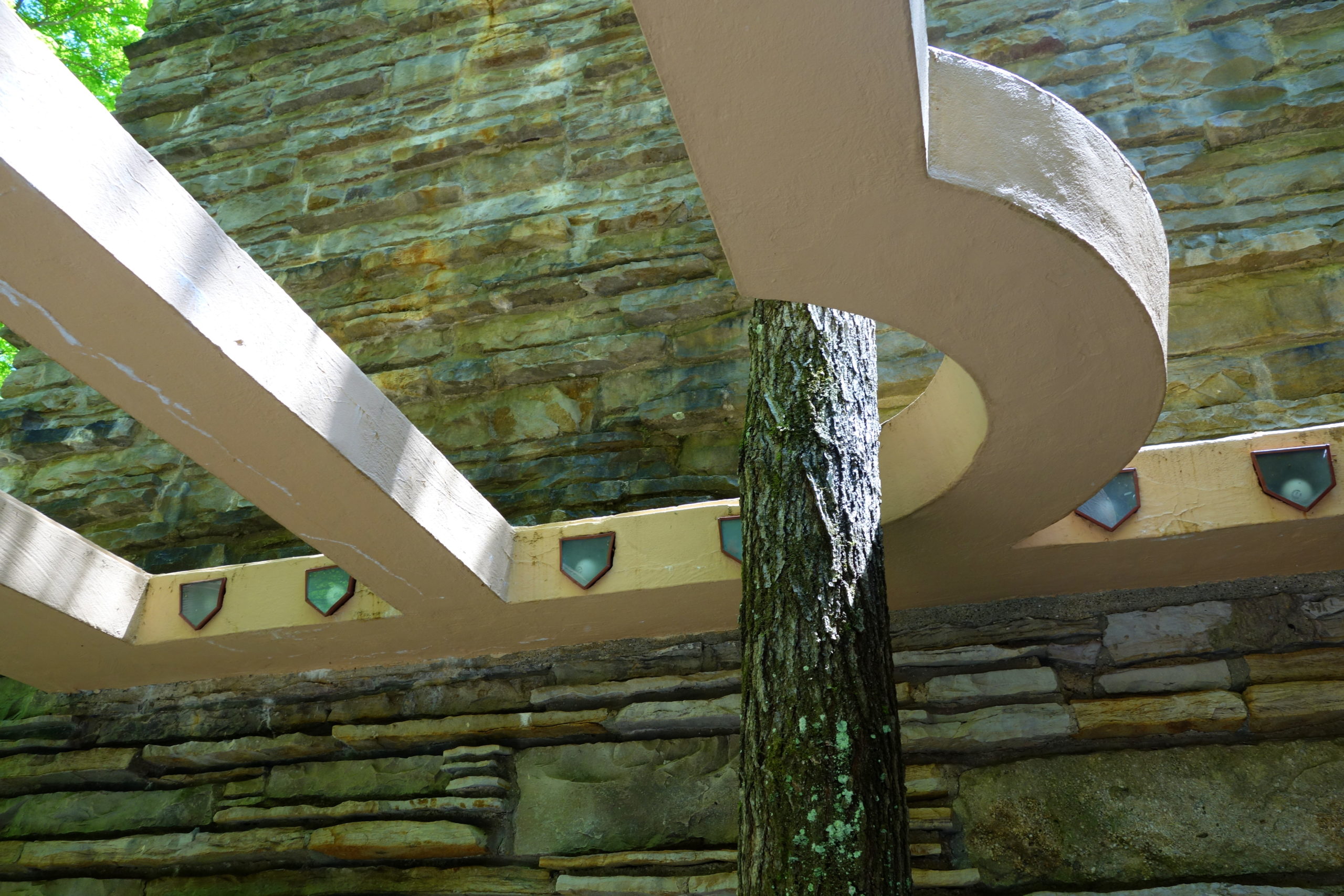 Photograph of an architectural structure featuring a curved beige concrete beam supported by a tree trunk against a stone wall background. The design highlights integration of natural elements with modern construction, with sunlight casting shadows on beams and textured stone surfaces.