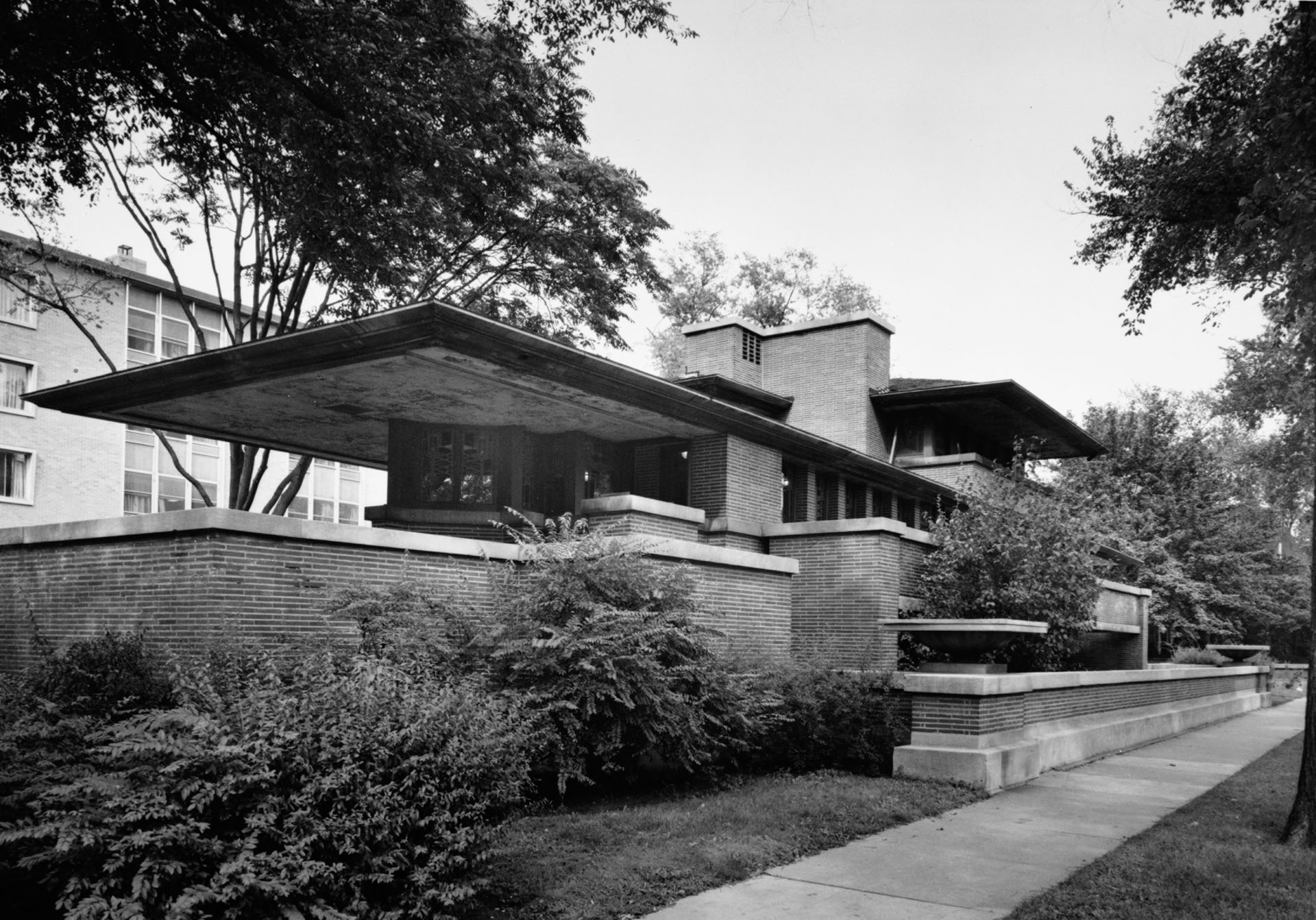 Black and white photograph of a Prairie-style house featuring strong horizontal lines, overhanging eaves, and extensive use of brick and wood. The structure is surrounded by dense shrubbery and trees, with a sidewalk running alongside the property.