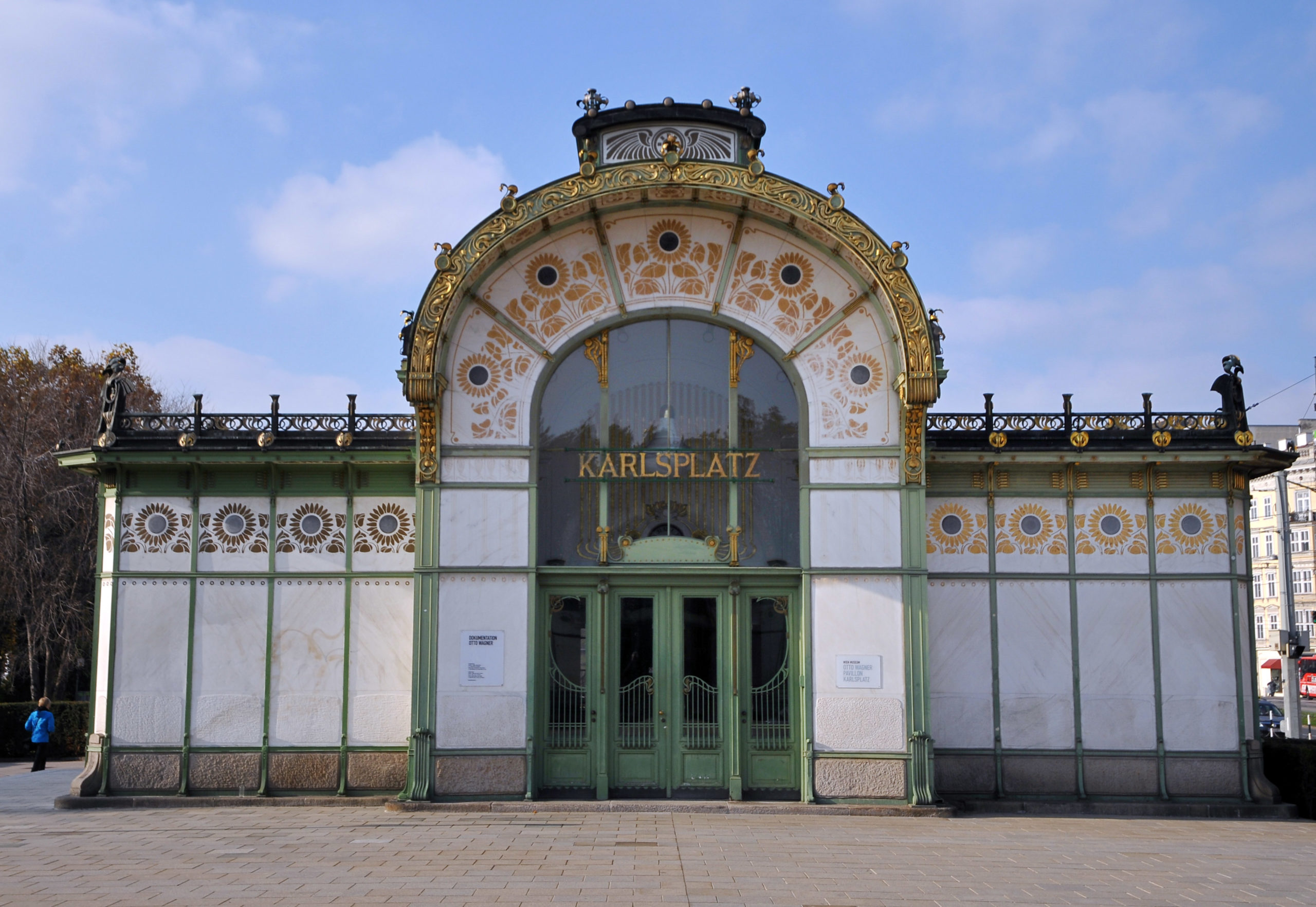 Photograph of Karlsplatz pavilion, a historic Art Nouveau building with a large arched window and ornate gold and green decorative elements. The structure features symmetrical design, floral patterns, and the name "KARLSPLATZ" displayed above green double doors.