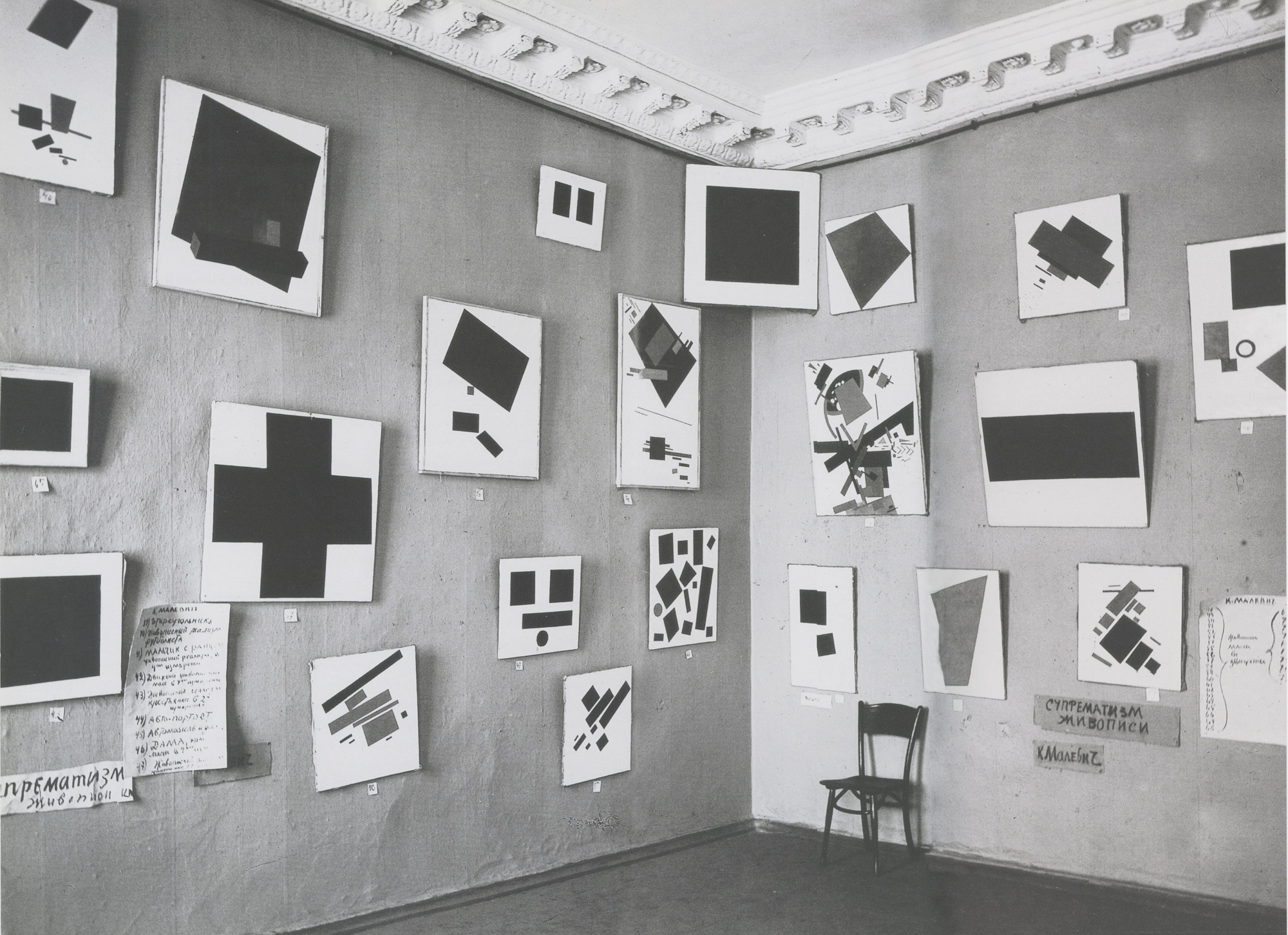 Black and white photograph of an art gallery corner displaying multiple abstract geometric paintings mounted on walls. Paintings feature bold shapes like squares, rectangles, and crosses in solid black on white backgrounds, with handwritten notes and a single chair adding context to the exhibition.