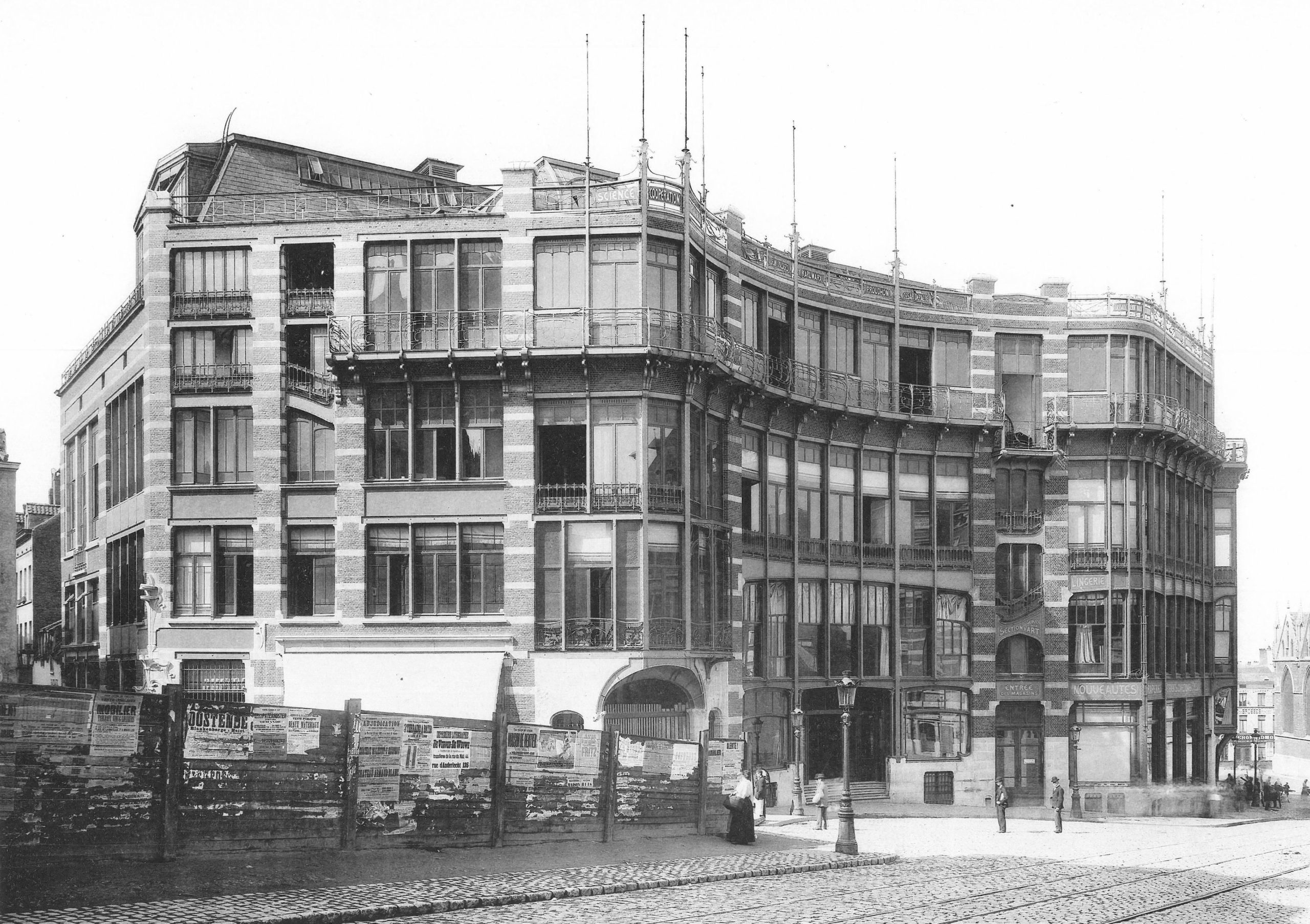 Black and white photograph of a large, multi-story building with extensive glass windows and ornate architectural details, situated on a cobblestone street corner. The structure features a mix of brick and stone materials, with visible scaffolding or poles on the roof, indicating possible construction or renovation.
