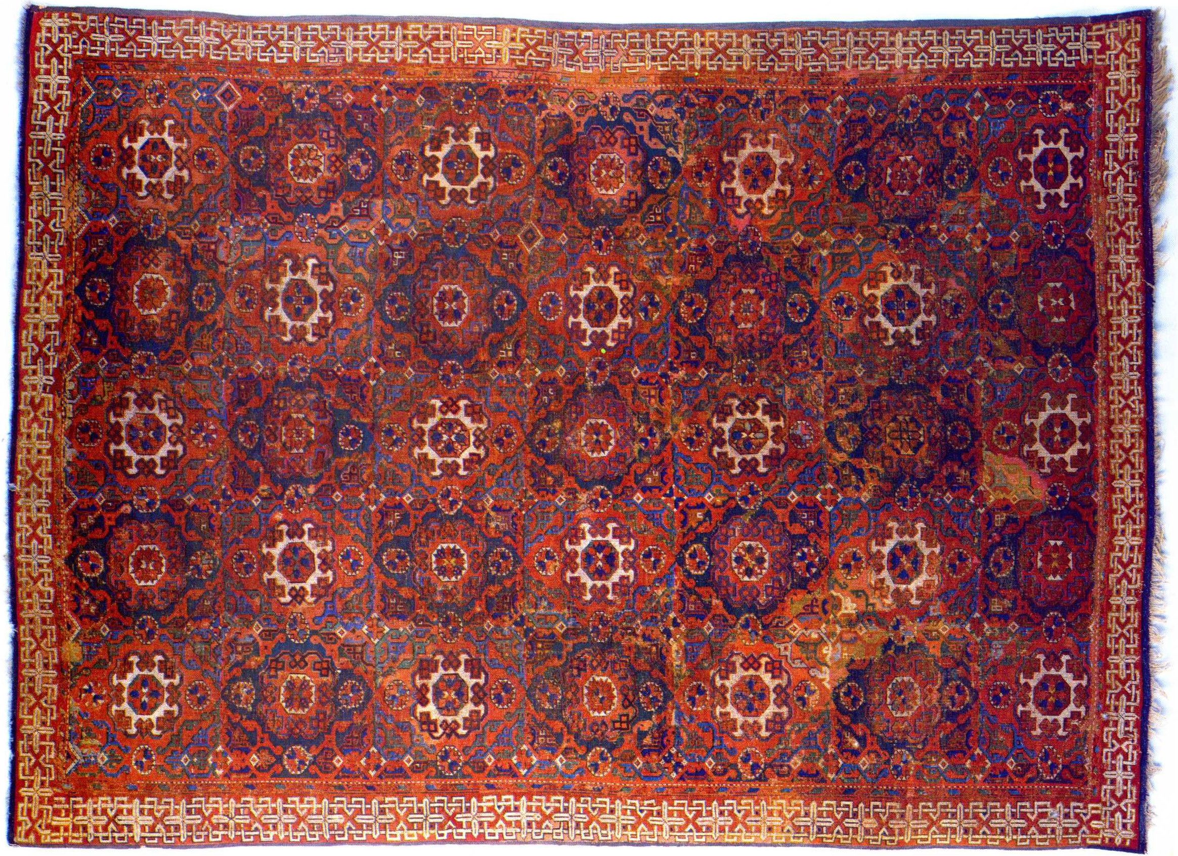 Photograph of a Holbein carpet, 15th–16th century, wool, from Turkey, featuring an intricate pattern of circular floral motifs in red, blue, and white tones. The rug has a decorative border with geometric designs and displays areas of wear and discoloration, indicating age and use.