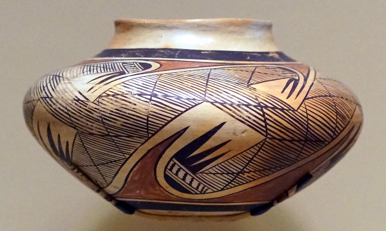 Photograph of a round ceramic pot with a wide body and narrow neck, featuring intricate geometric and abstract patterns in black and brown on a beige background. Design includes cross-hatching, curved lines, and sharp angular shapes, highlighting traditional craftsmanship and artistic style.