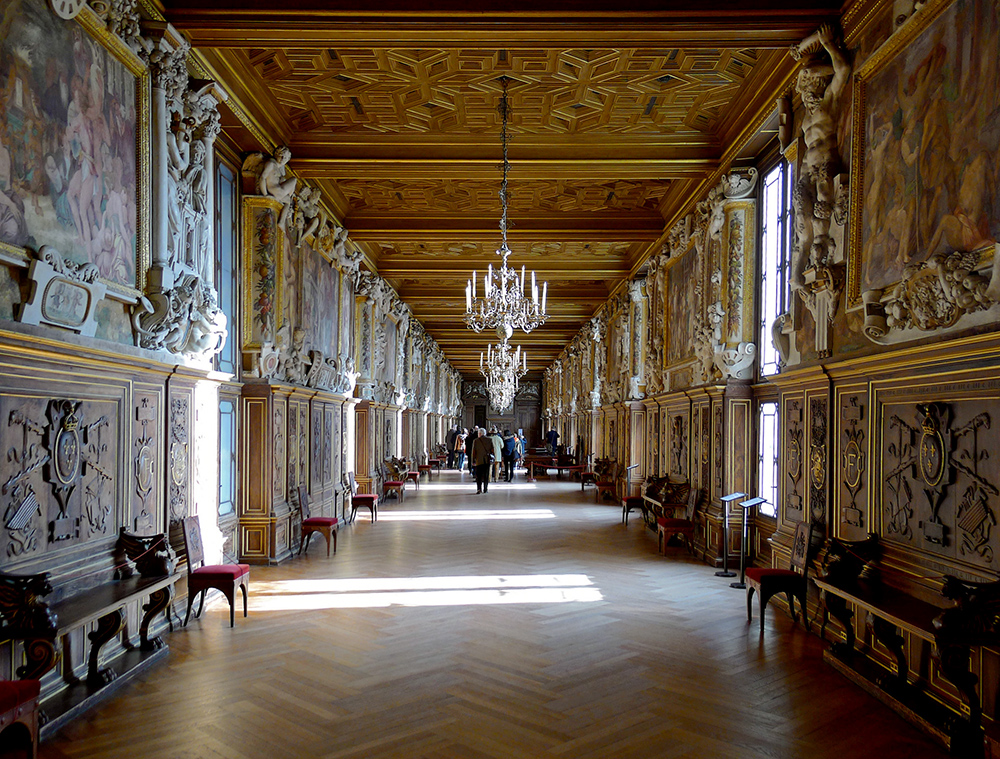 Photograph of an ornate, long hallway with wooden parquet flooring, intricate carved wooden paneling, and large paintings above the paneling on both sides. The ceiling features detailed woodwork and chandeliers, with natural light streaming through tall windows, highlighting the historical and artistic grandeur of the space.