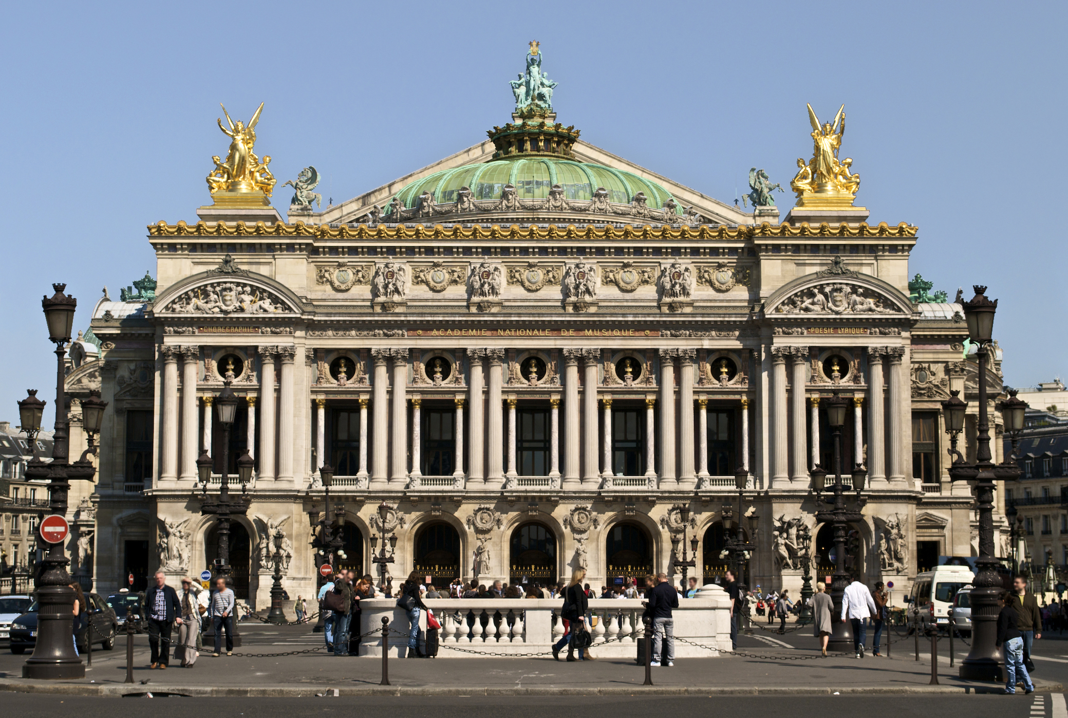 Photograph of Palais Garnier, a historic opera house in Paris, showcasing its ornate Beaux-Arts architecture with a green copper dome, golden statues, and detailed stone carvings. People walking and standing in front highlight its status as a popular cultural landmark and tourist attraction.