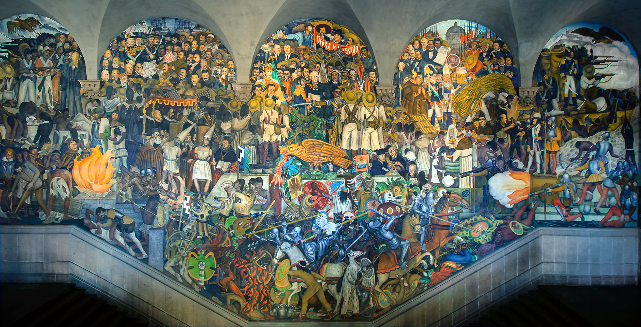 A mural depicting a vibrant historical scene with numerous figures engaged in various activities, including battles, celebrations, and cultural rituals. The artwork features rich colors, intricate details, and symbolic elements such as warriors, animals, and traditional attire, illustrating a complex narrative of heritage and conflict.