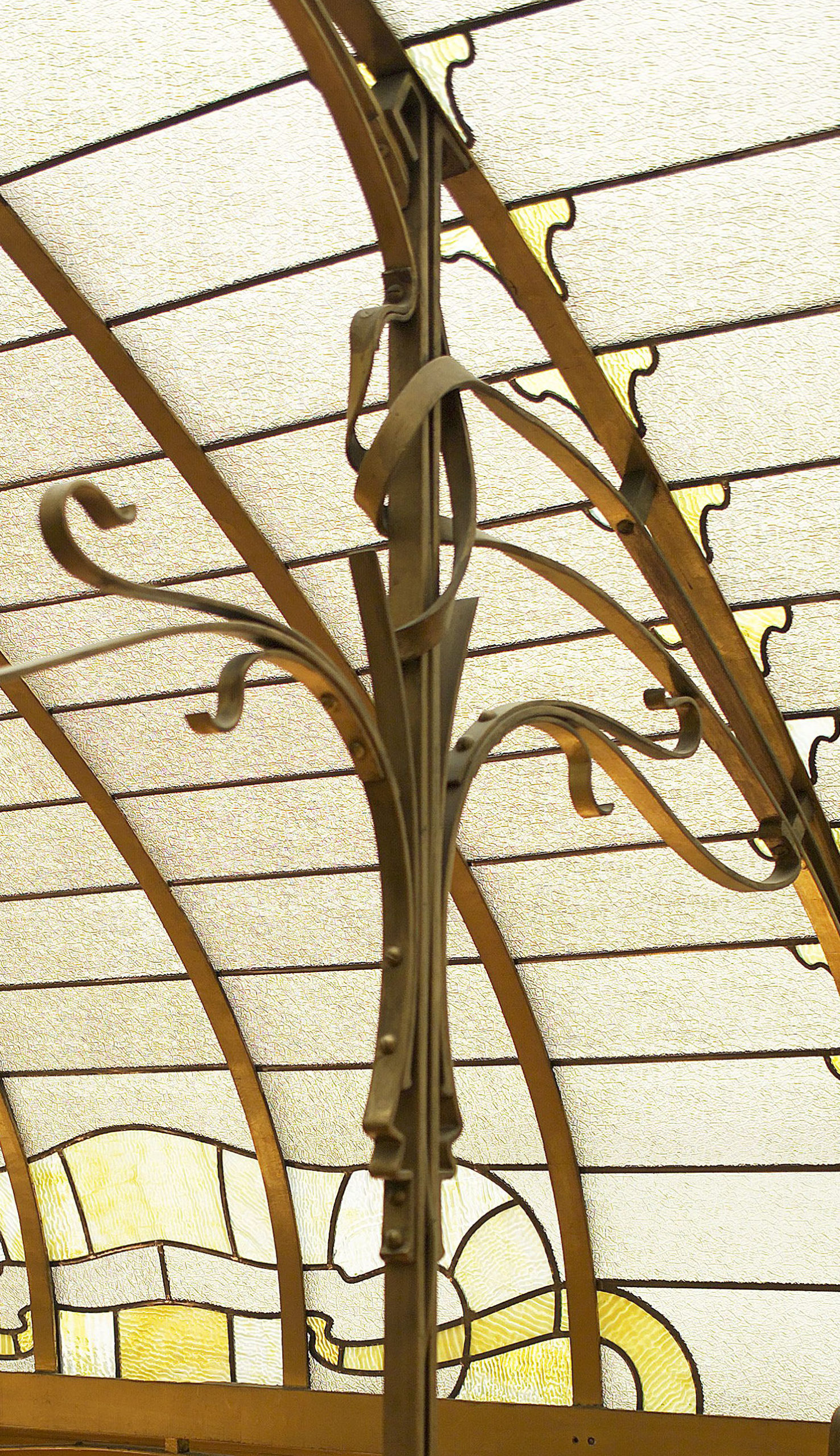 Photograph of an ornate wrought iron support structure holding up a translucent, segmented glass ceiling. The ceiling features curved yellow and white glass panels with decorative iron hooks and rivets along the metal framework.