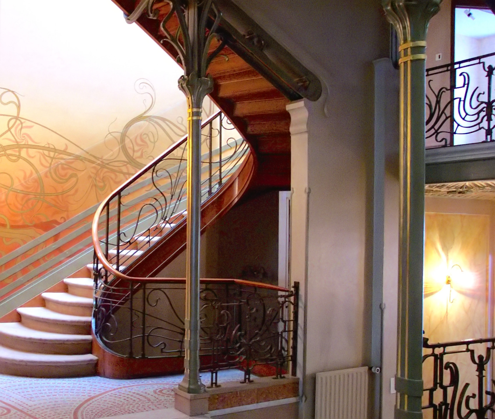 Photograph of an ornate spiral staircase inside a historic building, featuring intricate wrought iron railings and wooden steps. Warm lighting highlights decorative columns and patterned flooring, emphasizing architectural elegance and vintage design elements.