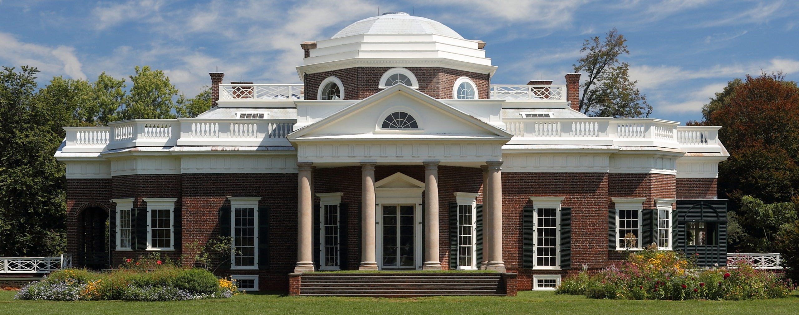 Photograph of Thomas Jefferson's Monticello neoclassical mansion with a prominent white dome, brick facade, and tall white columns at the entrance. The building is surrounded by green grass and trees under a partly cloudy blue sky.