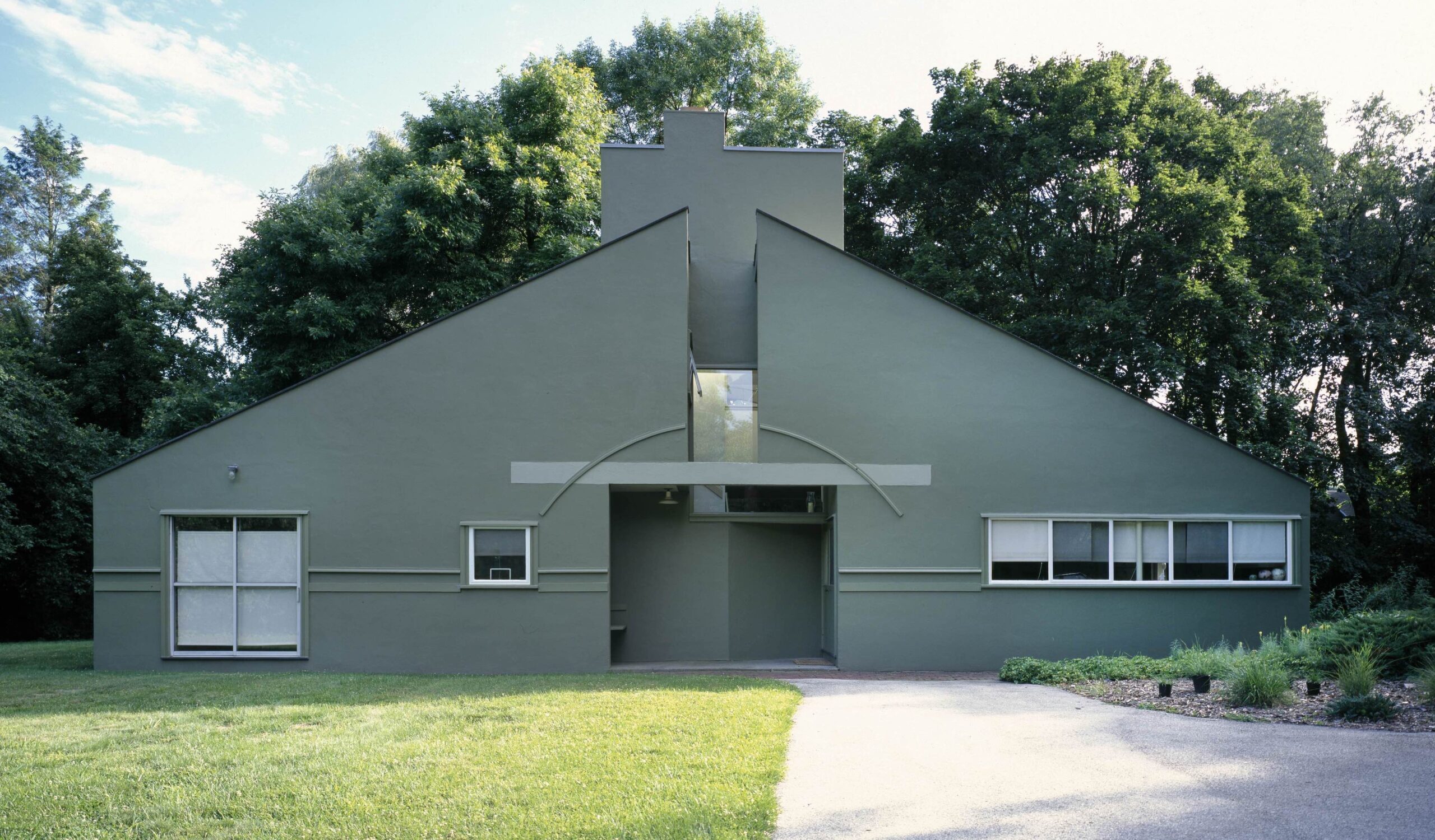 Photograph of a modern, symmetrical house with a flat roof and two large triangular walls painted gray, surrounded by green grass and trees. The house features a central entrance with a small overhang, multiple rectangular windows, and a prominent chimney-like structure at the top center.