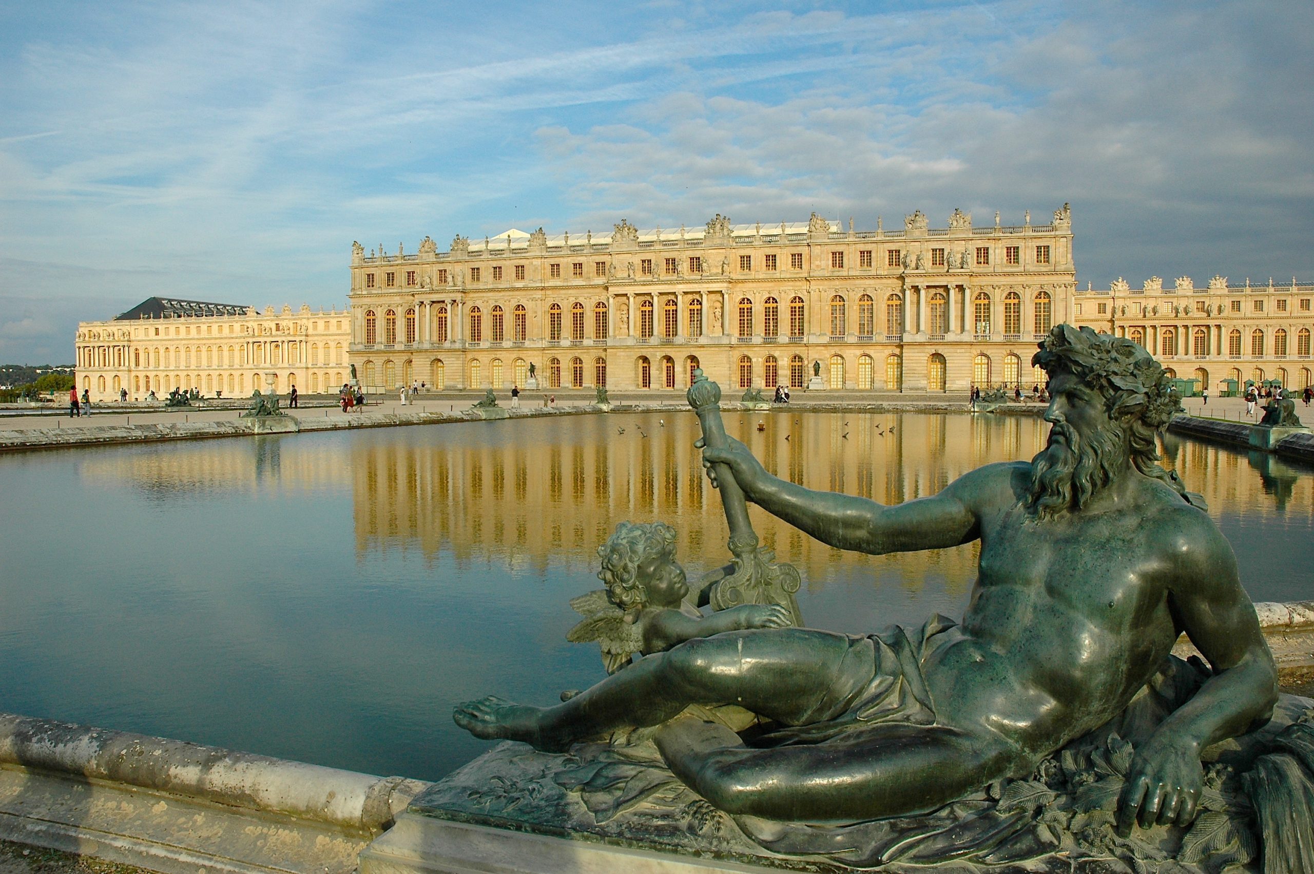 Photograph of Palace of Versailles with a large reflecting pool and bronze statue of a reclining male figure holding a staff in foreground. Golden sunlight illuminates palace facade and highlights detailed sculptures around pool under partly cloudy blue sky.