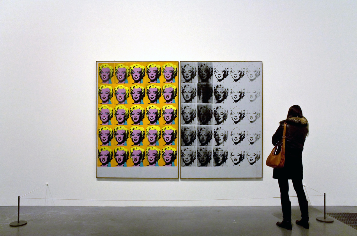 Photograph of a gallery setting featuring two adjacent pop art portraits of Marilyn Monroe, one in vibrant colors with yellow, pink, and blue tones, and the other in grayscale. A person stands observing the artwork, highlighting contrast between colorful repetition and monochrome fading effects, emphasizing themes of celebrity and media reproduction.