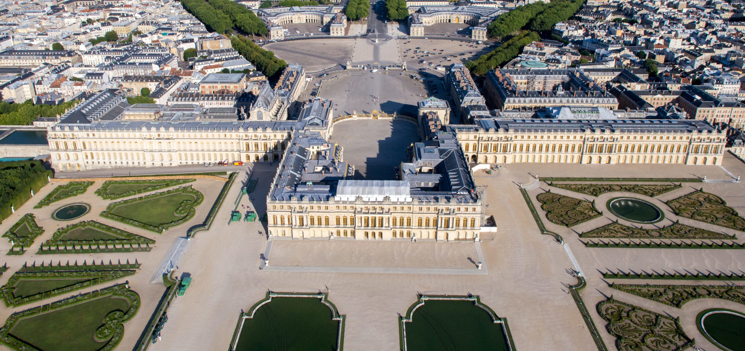Aerial photograph of the Palace of Versailles showcasing its symmetrical gardens, ornate fountains, and expansive courtyards. The image highlights the grand architecture, geometric landscaping, and the palace's central position within the historic estate.