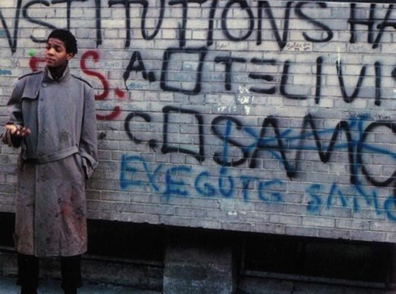 Photograph of a person standing against a graffiti-covered brick wall, wearing a long coat and looking to the side. Graffiti includes large black and blue letters with some red markings, featuring words and symbols that appear to be political or social statements.