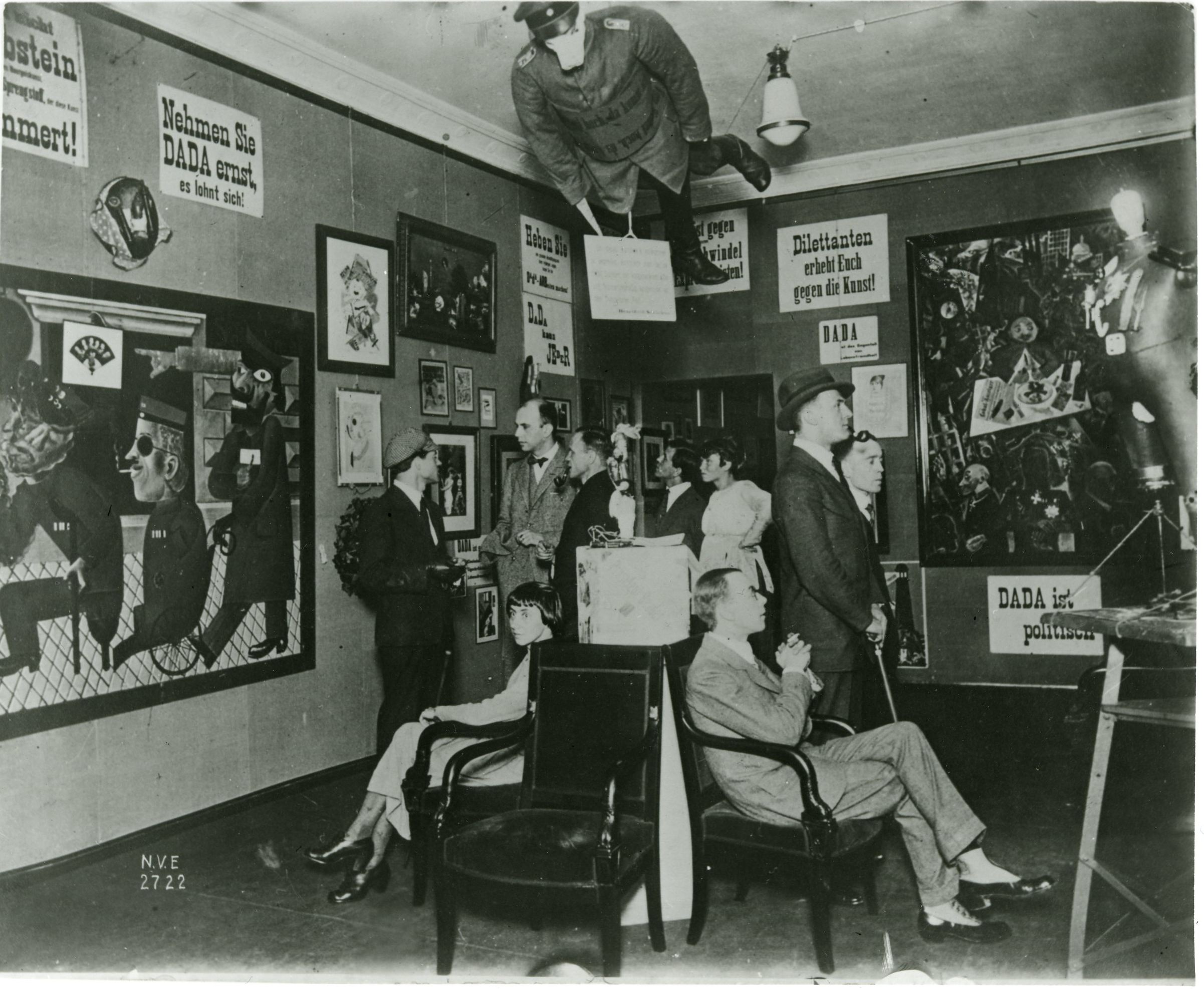 Black and white photograph of an art exhibition featuring Dada movement works, with several people observing paintings and posters on walls. Notable elements include a mannequin dressed as a soldier suspended from ceiling and various Dada slogans in German displayed prominently around the room.