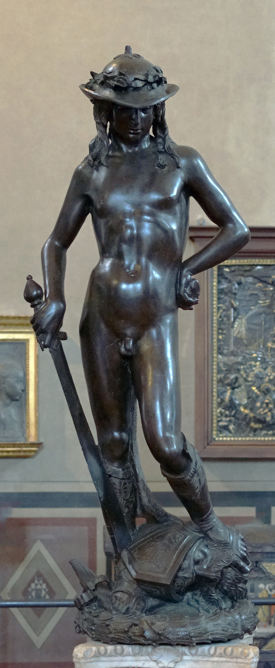 Donatello's David bronze sculpture of a young shepherd boy in the nude except for sandals and a hat. David stands on the head of goliath while holding a sword.