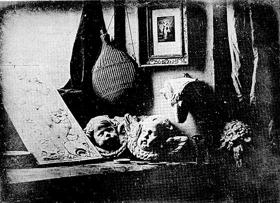 Black and white photograph of a tabletop displaying various sculpted masks and decorative objects, including a framed picture hanging on the wall. The scene highlights artistic craftsmanship with detailed facial features on masks and textured surfaces, suggesting a workshop or studio setting.