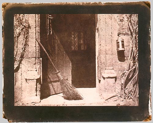 Sepia-toned photograph depicting an open doorway with a broom leaning against the doorframe, surrounded by textured stone walls and climbing vines. The composition highlights rustic architectural details and evokes a quiet, abandoned atmosphere.