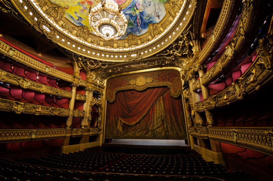 Photograph of an ornate theater interior featuring a grand stage with a richly decorated red curtain and multiple tiers of balconies adorned with gold accents and red seating. The ceiling showcases an elaborate chandelier and a colorful painting, highlighting the theater's luxurious and historic design.