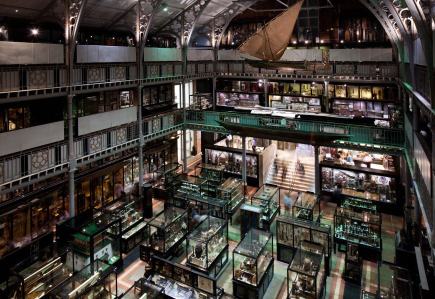 Photograph of a multi-level museum interior showcasing numerous glass display cases filled with artifacts and exhibits, with a large suspended sailboat model near the ceiling. The space features metal framework, balconies, and illuminated displays, highlighting historical and scientific collections.