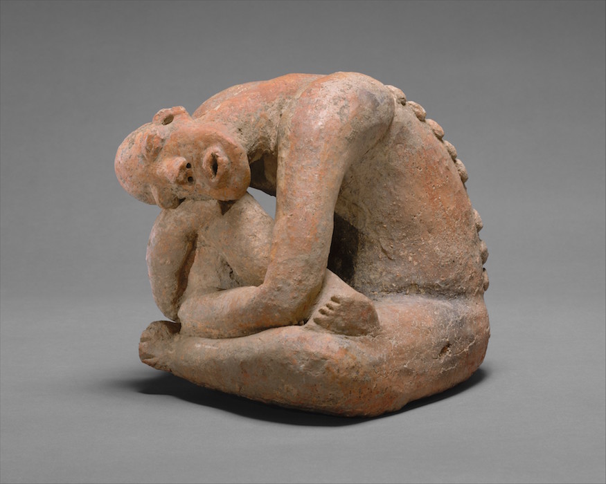 Photograph of a seated, curled human figure sculpture made of reddish-brown material with a textured surface. The figure has a contemplative expression, bent knees, and a series of small rounded protrusions along the spine.