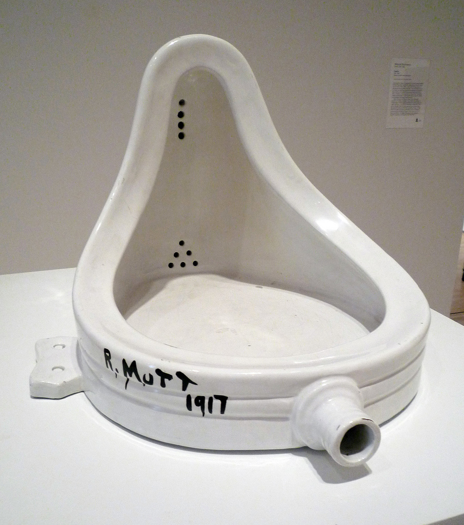Photograph of a white porcelain urinal titled "R. Mutt 1917," an iconic piece of modern art by Marcel Duchamp. The urinal is displayed on a white pedestal with black handwritten text on its front, highlighting its role in challenging traditional art concepts.