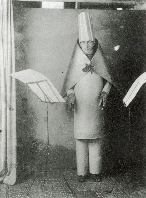 Black and white photograph of a person dressed in an unusual, cone-shaped costume with a tall, cylindrical hat, standing between two music stands holding sheet music. The setting appears to be a staged or theatrical environment, highlighting an eccentric or avant-garde fashion or performance art style.