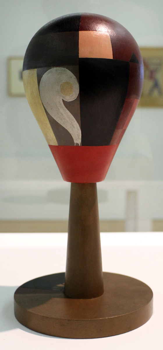 Photograph of a modernist sculpture resembling a maraca or rattle, featuring a rounded top with geometric color blocks in black, red, beige, and white, mounted on a cylindrical brown base. The design includes a prominent white swirl shape on one side, highlighting abstract artistic elements and contrasting color patterns.