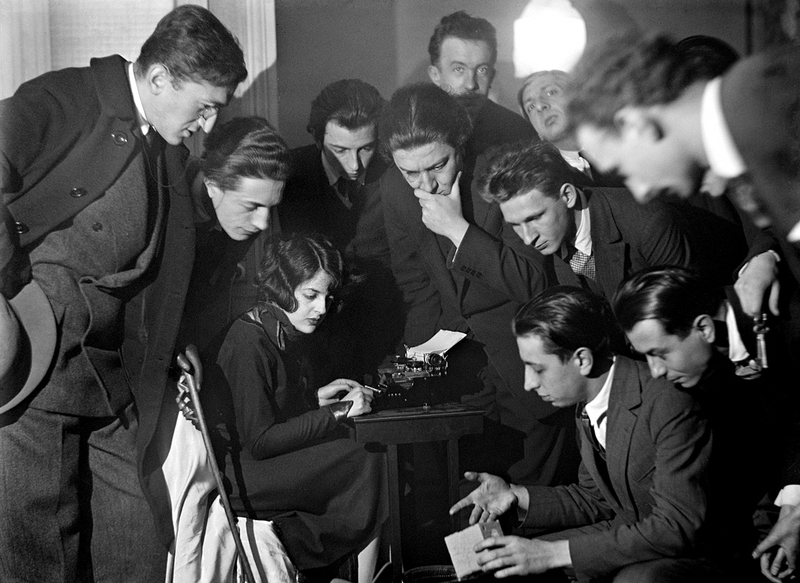 Black and white photograph showing a group of young men and one woman gathered closely around a small table with a typewriter, focused on the woman typing. The scene captures an intense moment of collaboration or instruction, with individuals displaying curiosity and concentration, dressed in early 20th-century formal attire.
