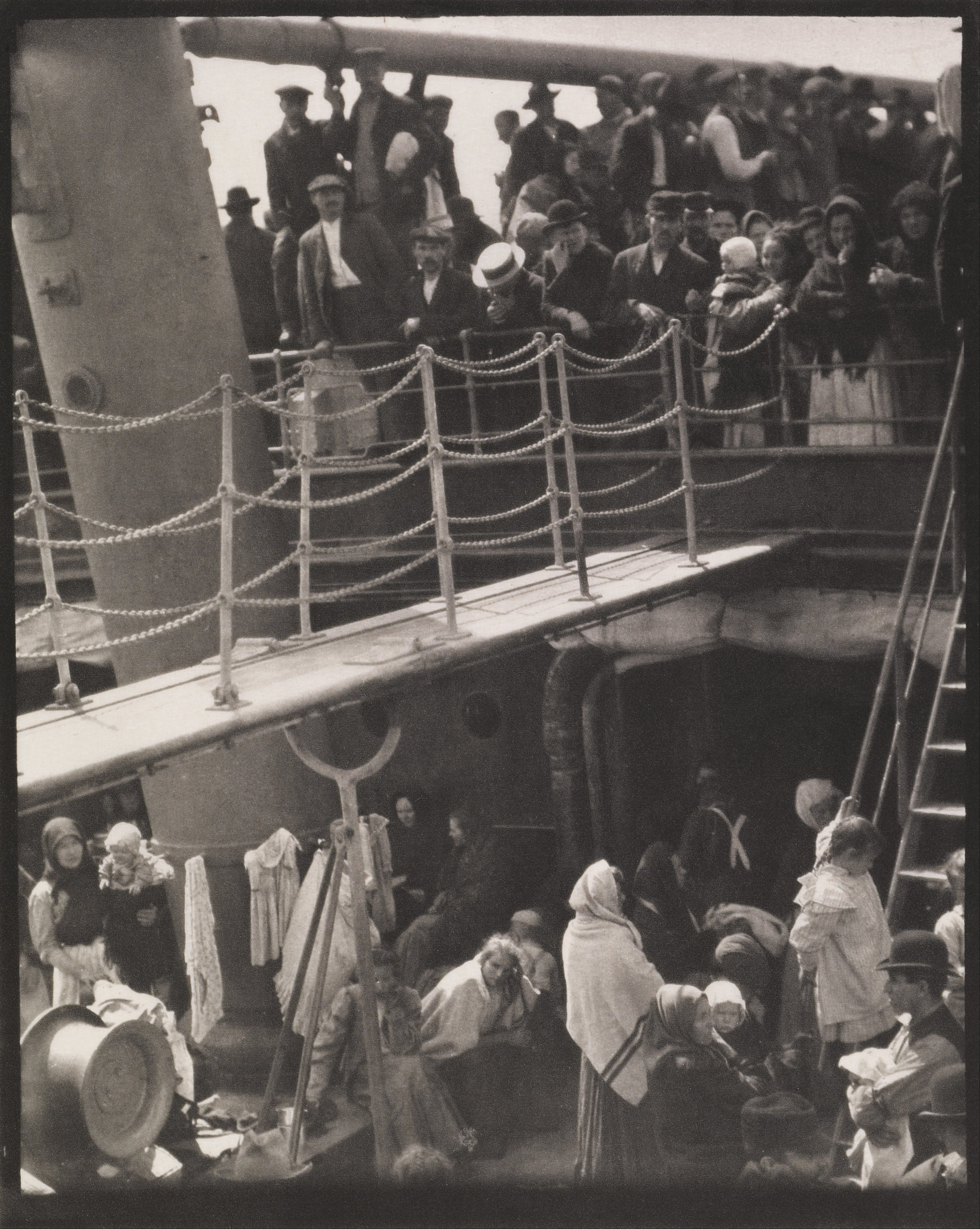 Black and white photograph showing a crowded ship deck with men, women, and children dressed in early 20th-century clothing. The scene captures passengers standing and sitting near ropes and ship structures, highlighting a moment of travel or migration.