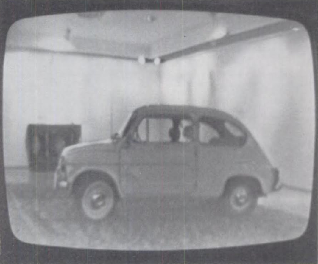 Black and white photograph of a vintage compact car displayed inside a minimalist room with plain walls and ceiling lights. The car is positioned sideways, showing its rounded shape and small size, with a slightly grainy texture suggesting an older or retro style.