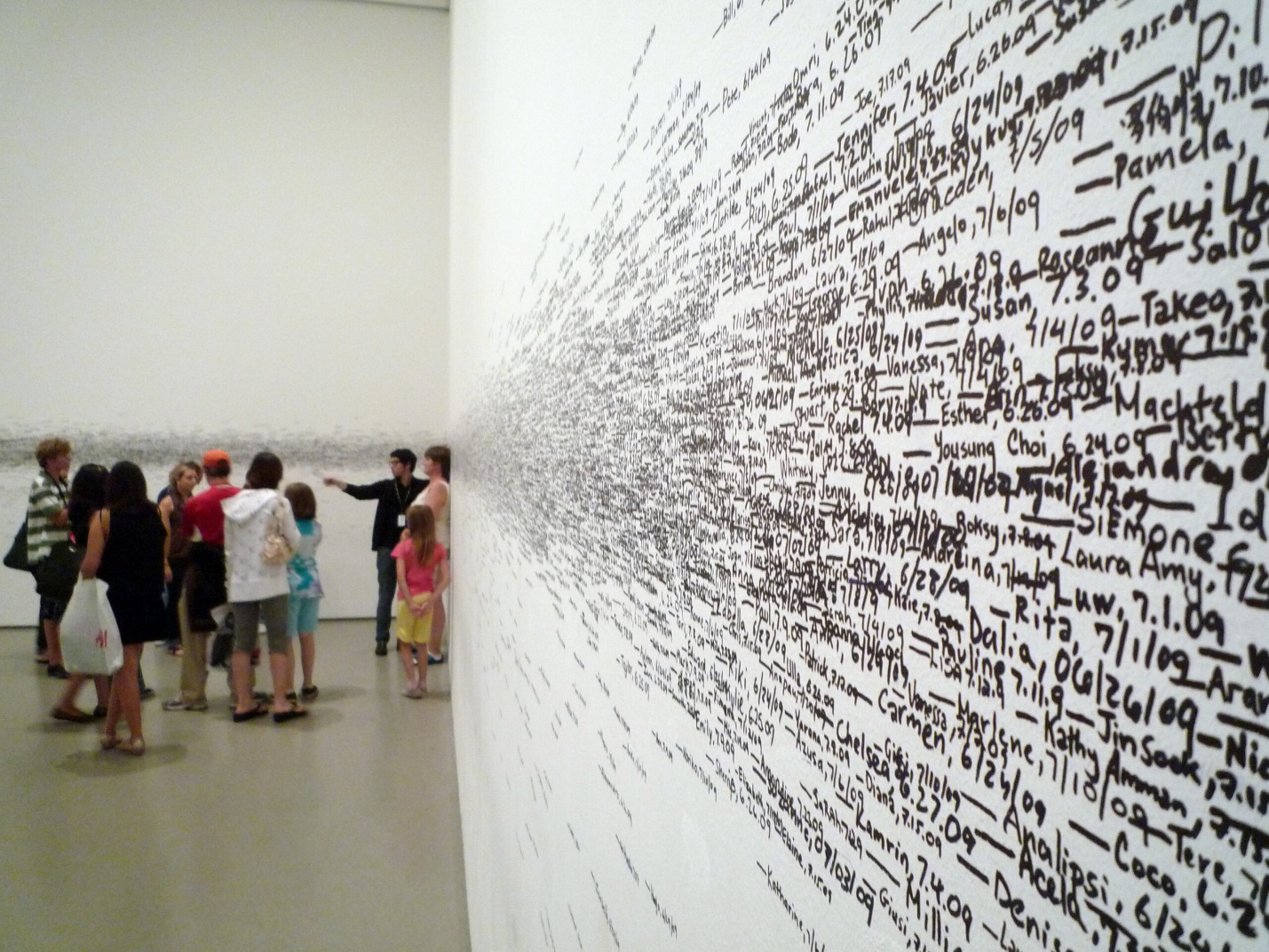 Photograph of a group of people observing a large wall covered in densely packed handwritten names and dates in black ink, arranged to create a perspective effect that draws the eye toward a vanishing point. The artwork emphasizes human presence and passage of time through the extensive list of names, with the crowd adding scale and engagement to the installation.
