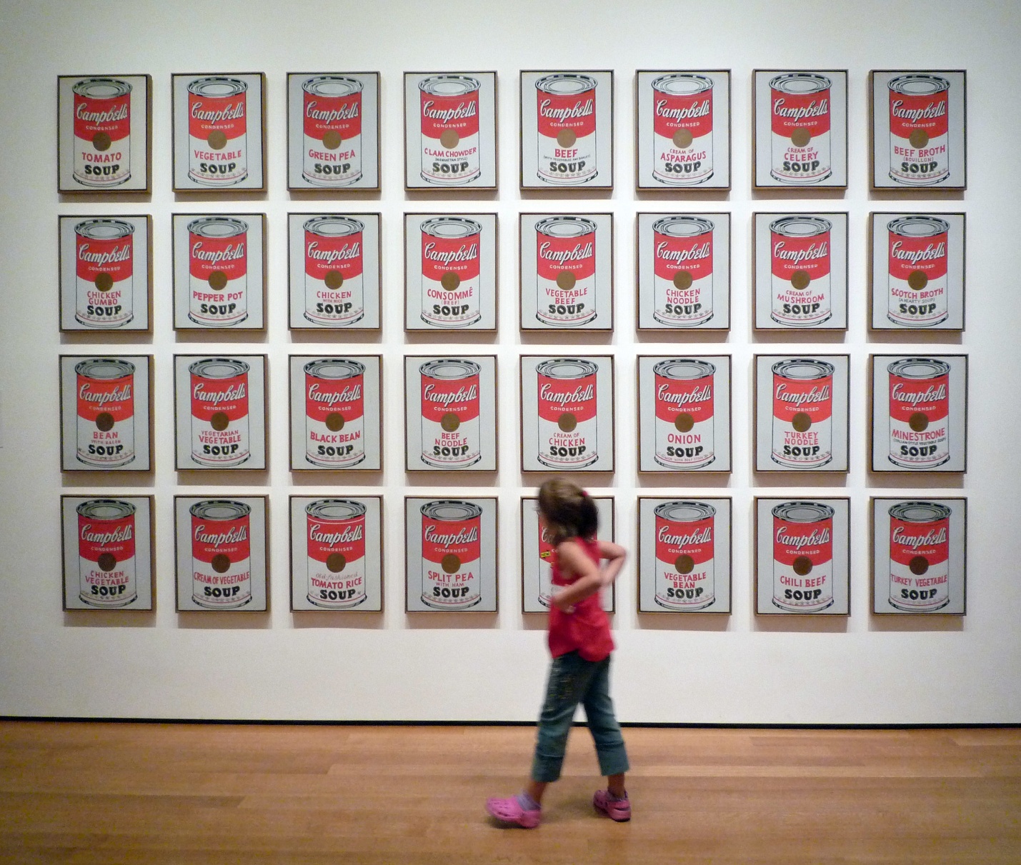 Photograph of a gallery wall displaying 32 framed Campbell's soup can prints arranged in four rows and eight columns, showcasing various soup flavors with red and white labels. A child in a red shirt and blue jeans walks in front, highlighting scale and interaction with iconic pop art pieces.