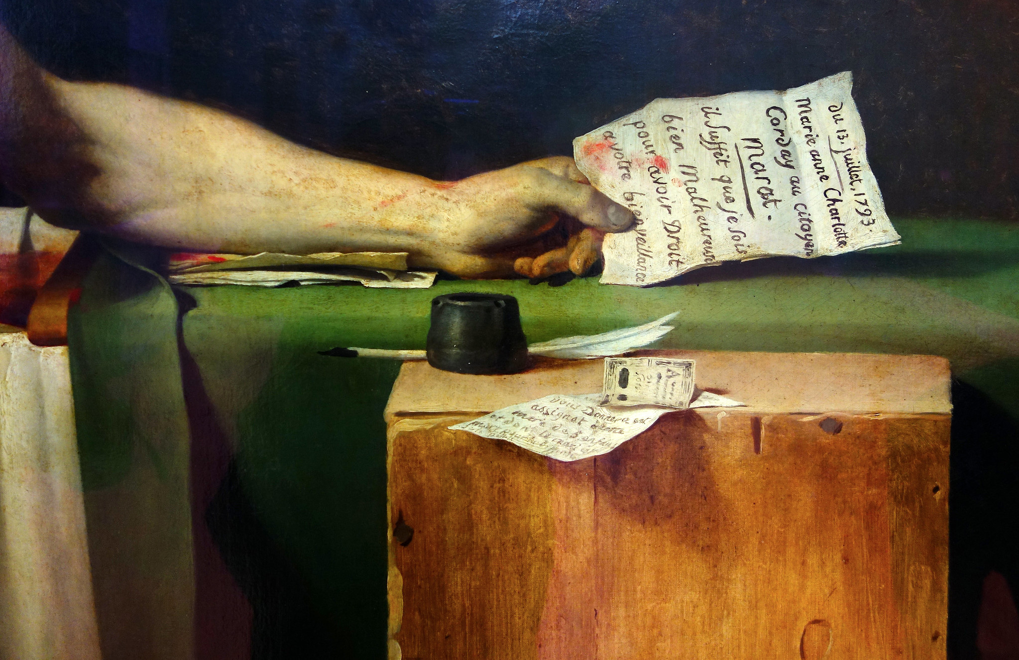 Detail of David's Death of Marat painting depicting a close-up of a person's arm resting on a green cloth-covered table, holding a handwritten note. The scene includes an inkwell, quill, and additional papers on a wooden box.