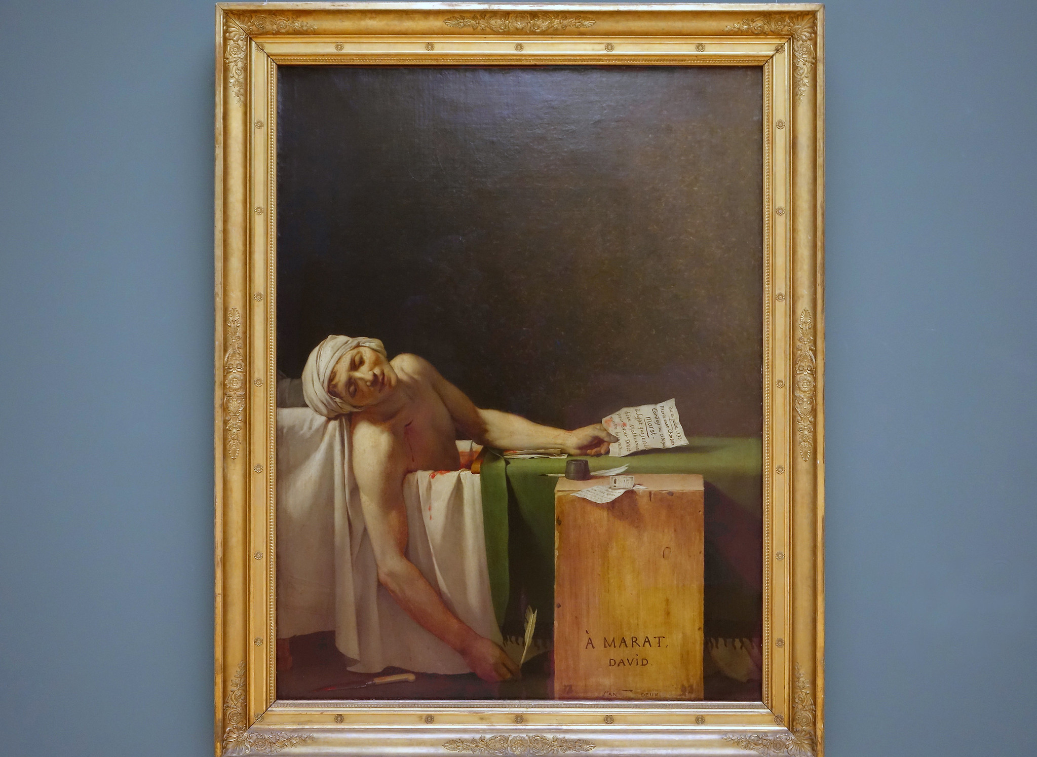 Jacques-Louis David, Death of Marat painting features a deceased figure reclining in a bathtub holding a quill in one hand a letter in the other. Next to the figure and tub is a wood writing table labeled with "A Marat." and signed "David"