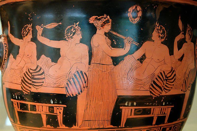 Painting of a symposium on a clay vessel featuring a woman slave entertainer and male guests reclining and drinking.