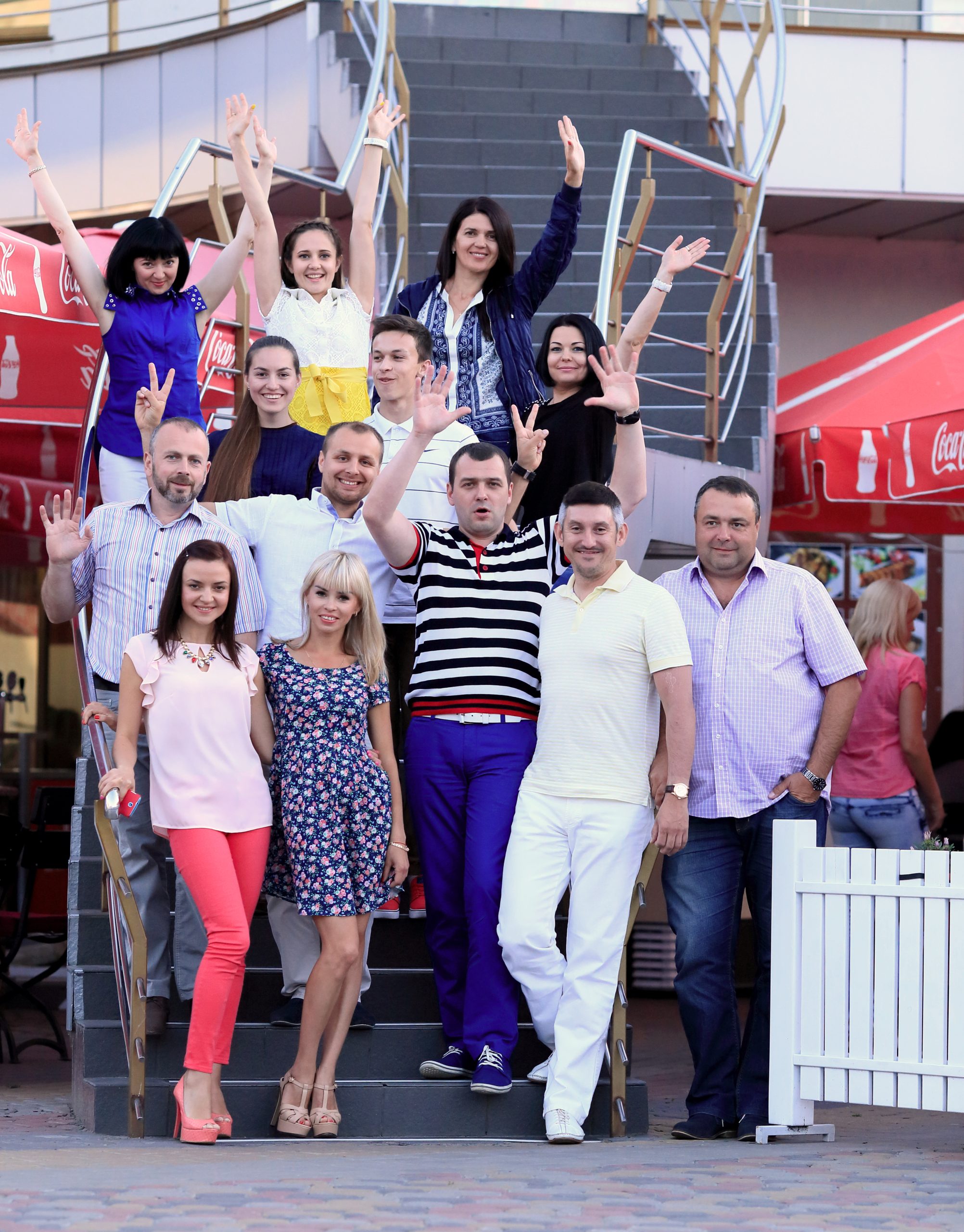 A group of people in colorful clothes stand together on a stairway. Some have their arms up, and most are smiling and laughing.