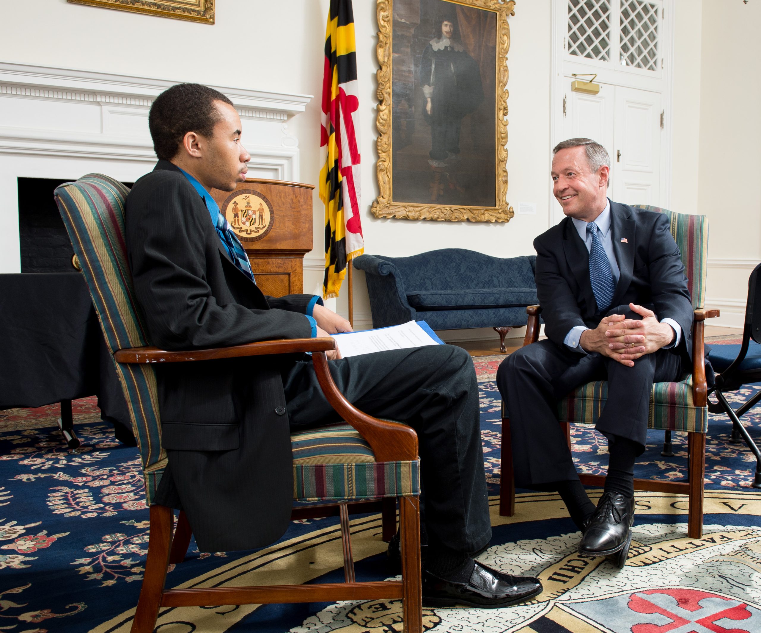 Two people sit in armchairs facing each other in a nice office. One is a younger male wearing a suit and holding a notepad in his lap. The person facing him is also wearing a dark suit while smiling with his hands on his knee.